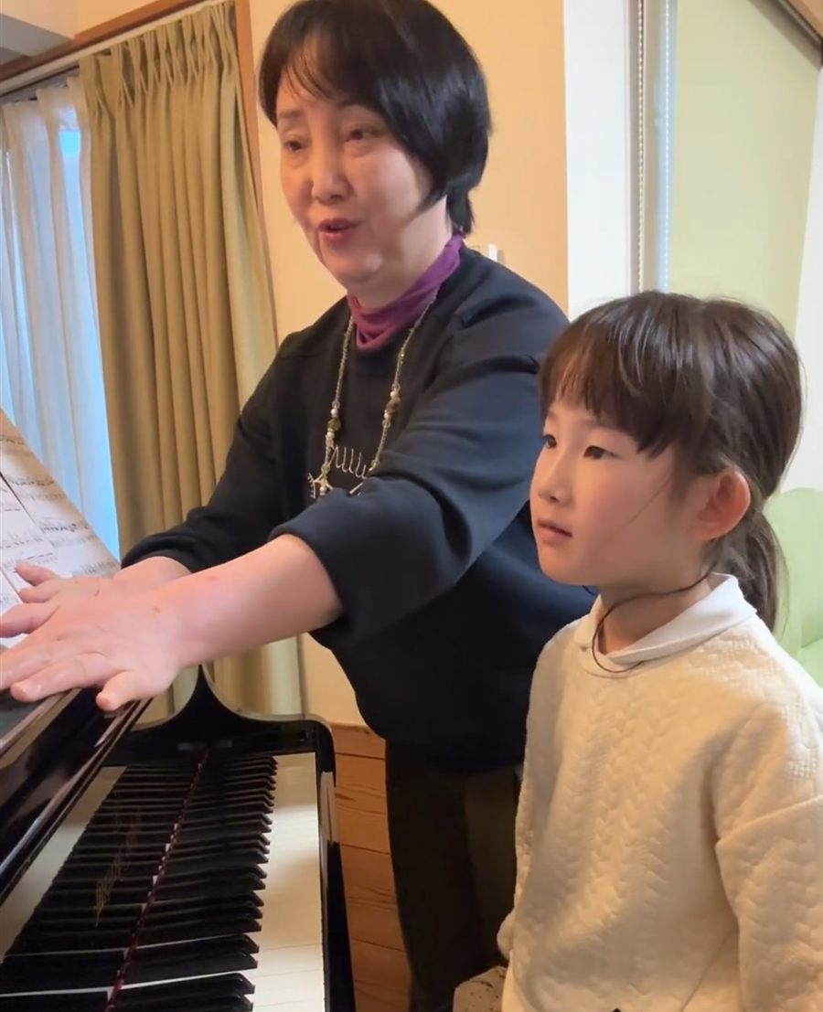 教室のグランドピアノで、講師がお手本を弾いて見せ、隣で女の子が見ているレッスン風景